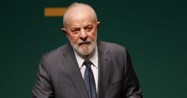 Pessoa usando blazer preto sobre camisa branca, complementada com um lenço cinza no pescoço. Ela está de braços cruzados diante de um fundo branco liso, transmitindo postura profissional e confiante.