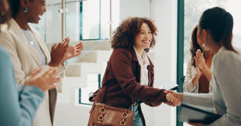 Mulher sorridente a apertar a mão a outra mulher num ambiente de escritório de Tecnologia, enquanto outras colegas aplaudem ao fundo, demonstrando boas-vindas ou celebração profissional.
