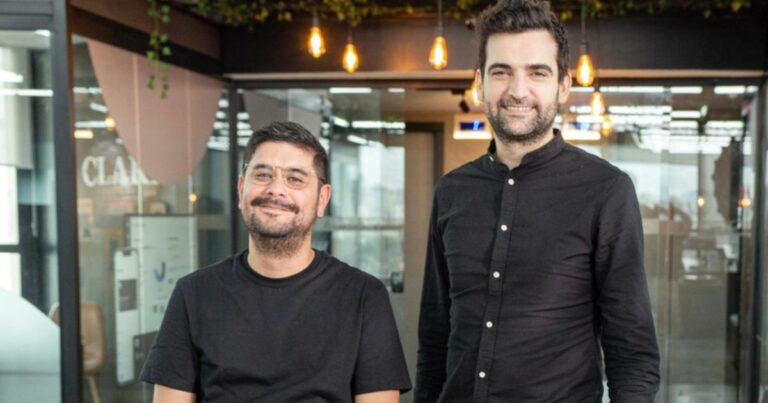 Dois homens a sorrir posam num ambiente de escritório moderno e iluminado. O homem à esquerda está sentado, usa óculos e uma t-shirt preta. O homem à direita está de pé, veste uma camisa preta de manga comprida. Ao fundo vê-se uma parede de vidro com iluminação suspensa e detalhes de plantas decorativas no teto.
