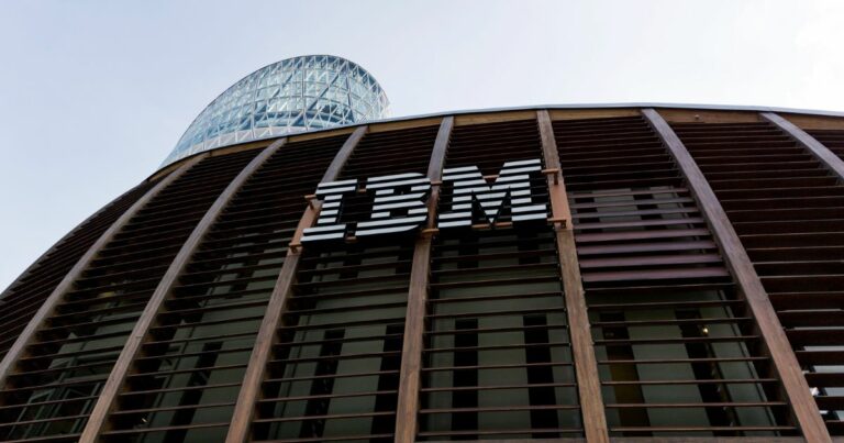 Fachada moderna de um edifício com o logótipo da IBM em destaque, com estrutura em madeira e vidro e uma torre envidraçada ao fundo."