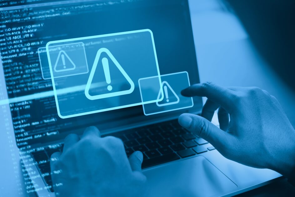 Imagem de um laptop com alertas de segurança e mensagens de erro na tela, indicando possíveis riscos de segurança digital ou vírus. A mão do usuário interagindo com o computador reforça o foco em cibersegurança e proteção de dados