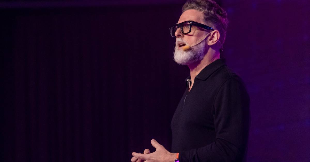 futuro: Neil Redding, CEO da Redding Futures e futurista, aparece em um palco durante uma palestra. Ele está de perfil, usando óculos de armação preta, microfone de cabeça e camisa preta de manga longa. O fundo é iluminado com luz roxa, criando um clima moderno e tecnológico.