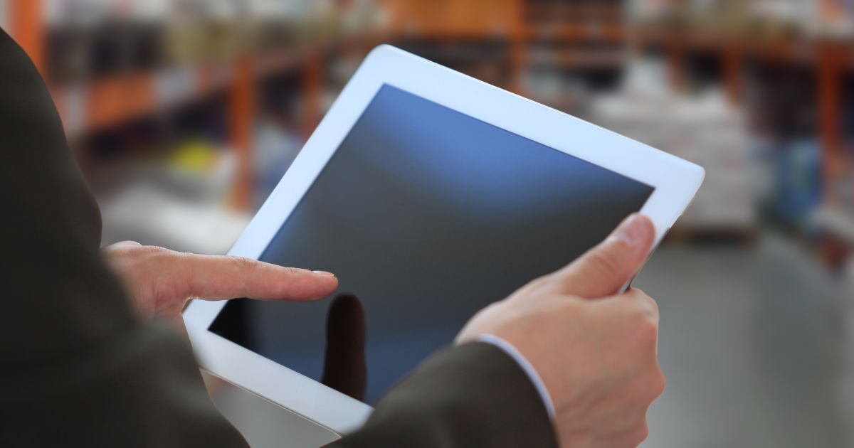 Uma pessoa segura um tablet branco com as duas mãos, enquanto toca a tela com o dedo indicador da mão direita. Ao fundo, o ambiente está desfocado, sugerindo um local interno como uma loja ou armazém, com prateleiras e diversos itens visíveis. (M.Dias Branco)