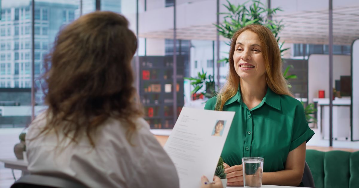 Duas mulheres estão em uma entrevista de emprego em um escritório moderno e iluminado. No primeiro plano, vemos a entrevistadora de costas, com cabelo castanho cacheado e vestindo uma blusa branca. Ela segura um currículo com a foto da candidata. A candidata, à frente, sorri enquanto fala; ela tem cabelos castanhos claros e lisos, veste uma camisa verde e está sentada à mesa. Na mesa há um copo de água e uma pequena planta decorativa. Ao fundo, grandes janelas de vidro mostram um ambiente corporativo com plantas e móveis modernos.