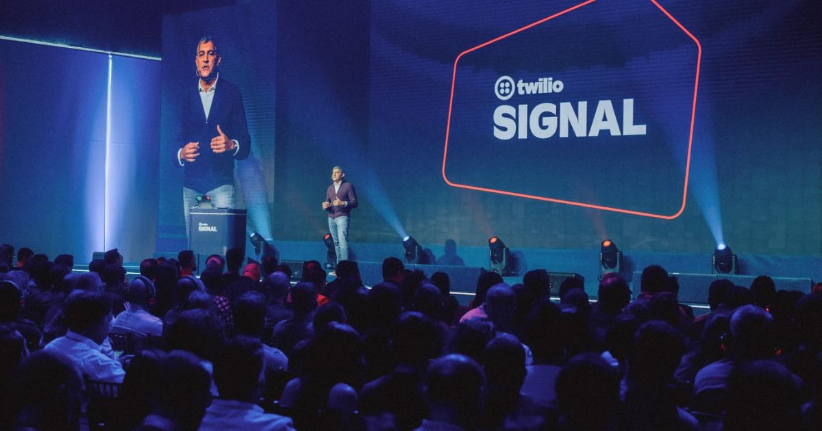 Um palestrante está no palco durante uma conferência ou evento, com o público sentado à frente. No fundo do palco, há uma grande tela exibindo o texto "twilio SIGNAL" junto com o logotipo da Twilio. À esquerda, uma tela menor mostra um close do palestrante.