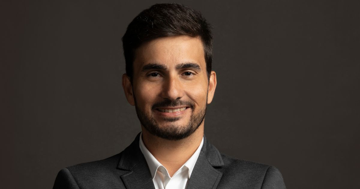 Homem jovem, de cabelos castanhos escuros e curtos, com barba bem aparada. Ele sorri levemente, veste um terno cinza escuro com camisa branca, e está posicionado contra um fundo liso, de tom escuro. Sua expressão transmite confiança e simpatia. (Intelbras)