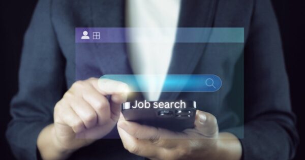 Uma pessoa vestindo um blazer escuro utiliza um smartphone. Sobre a imagem, há uma sobreposição gráfica de uma barra de pesquisa com o texto "Job search" digitado, sugerindo que a pessoa está procurando por vagas de emprego ou oportunidades profissionais. (tecnologia)