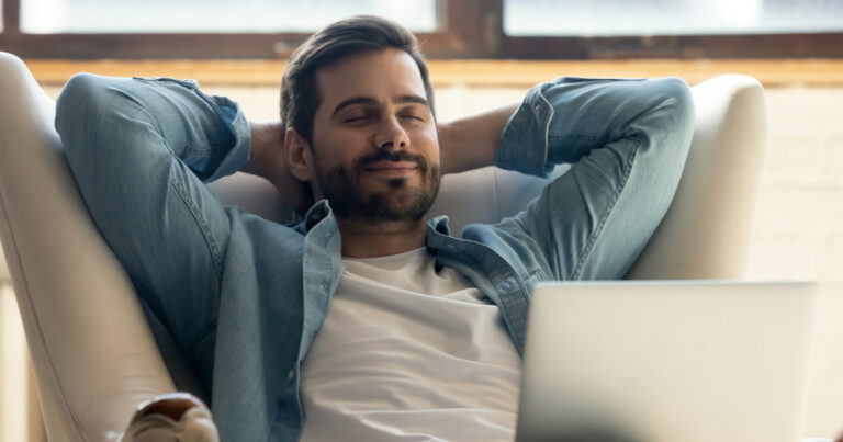 Homem jovem de barba curta, vestindo camiseta branca e camisa jeans aberta, sentado em uma poltrona clara com as mãos atrás da cabeça e expressão de relaxamento. À sua frente, há um notebook aberto, sugerindo que ele está aproveitando um momento de descanso em casa, possivelmente durante as férias. Ao fundo, há uma janela com luz natural entrando no ambiente (férias)