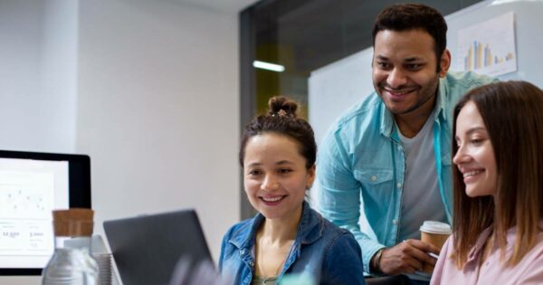Descrição da imagem para campo "Texto alternativo" do WordPress: Três jovens profissionais sorrindo e olhando para a tela de um laptop em um ambiente de escritório moderno. Ao fundo, há uma parede de vidro e um quadro branco com gráficos impressos. Um homem de camisa azul clara segura um copo de café enquanto interage com duas mulheres, uma de camisa jeans e outra de blusa rosa claro. Um monitor com gráficos de dados também é parcialmente visível à esquerda da imagem. Contexto: Porto Digital.
