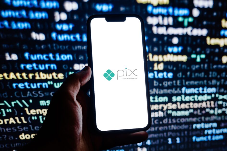 Pessoa segurando um smartphone exibindo a tela com o logo da plataforma PIX, enquanto o fundo mostra linhas de código em cores coloridas, destacando o tema de tecnologia e pagamentos digitais no Brasil