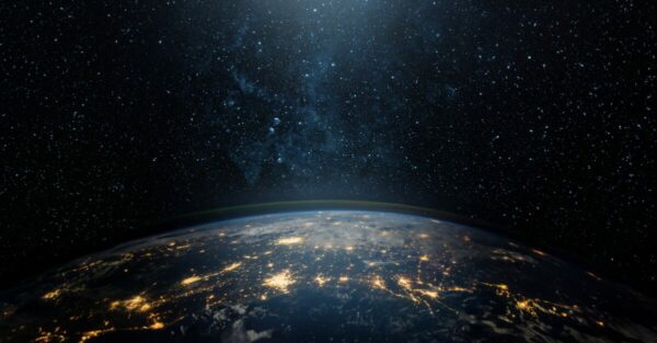 Imagem da Terra vista do espaço durante a noite, com a curvatura do planeta visível e diversas áreas iluminadas por luzes artificiais, representando grandes centros urbanos. Ao fundo, o céu escuro está repleto de estrelas, simbolizando a imensidão do universo e o conceito de globalização e conectividade mundial.