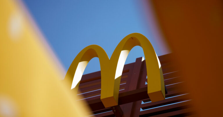Imagem com enquadramento artístico do logotipo do McDonald's, representado pelos arcos dourados em forma de "M", posicionado sobre uma estrutura de ripas de madeira, com o céu azul ao fundo. Partes desfocadas em amarelo e laranja nas bordas sugerem elementos em primeiro plano, adicionando profundidade à composição.