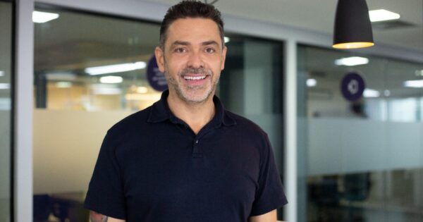 Uma pessoa, identificada como Julio Duram, CTO da Certisign, está em pé em um ambiente interno com um sorriso no rosto. Ele veste uma camisa polo escura. Ao fundo, há um escritório com paredes e portas de vidro. Algumas portas têm sinais circulares. As luzes do teto estão acesas e há reflexos visíveis nas superfícies de vidro.