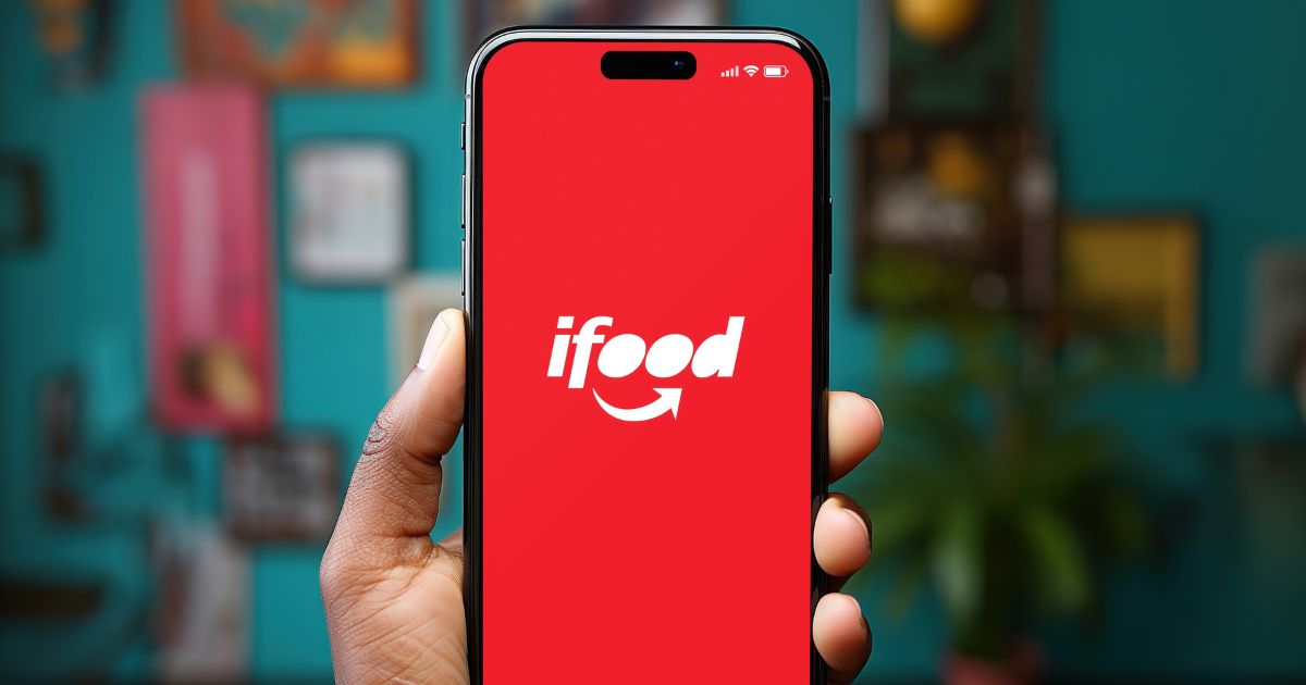Uma mão segura um smartphone com o aplicativo iFood aberto na tela. O visor exibe o logotipo do iFood, composto pela palavra "iFood" em texto branco sobre um fundo vermelho, com uma seta estilizada em forma de sorriso abaixo. O fundo da imagem está desfocado, mas é possível identificar um ambiente interno com quadros emoldurados e uma planta.