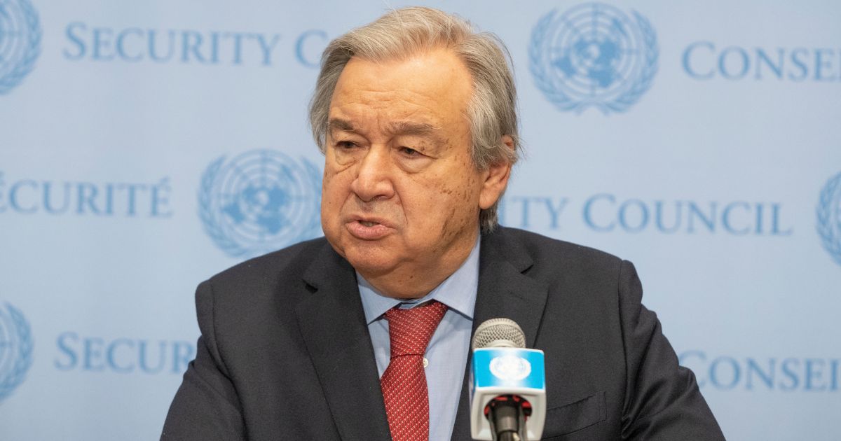Uma pessoa vestida formalmente com terno escuro, camisa azul clara e gravata vermelha está posicionada diante de um microfone que exibe o logotipo da ONU. Ao fundo, vê-se um painel azul claro com os dizeres "SECURITY COUNCIL" e "CONSEIL DE SÉCURITÉ", acompanhados por múltiplas repetições do emblema das Nações Unidas.
