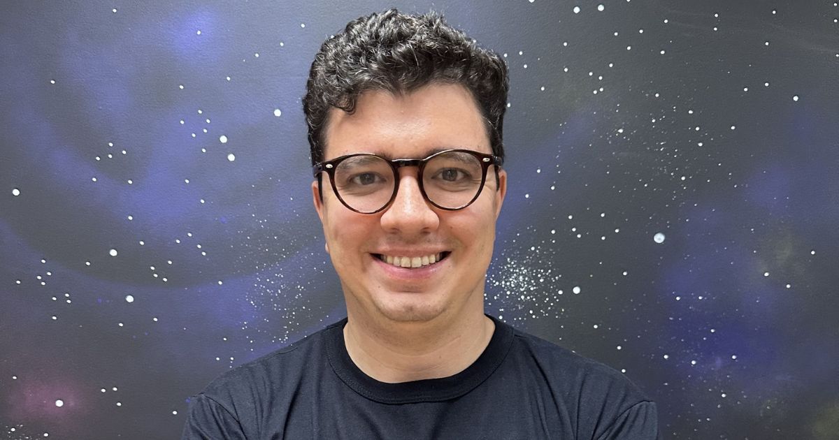 Uma pessoa com o rosto desfocado está em pé diante de um fundo que lembra o espaço sideral, com estrelas e nebulosas visíveis. A pessoa veste uma camiseta preta. (varejo)
