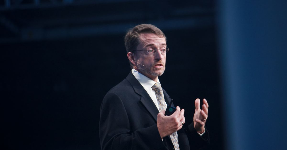 Pat Gelsinger, ex-CEO da Intel, vestido com terno e gravata está em pé, gesticulando com ambas as mãos. O rosto da pessoa está desfocado. O fundo é escuro, sugerindo um ambiente interno, possivelmente durante uma apresentação ou discurso. (Intel)