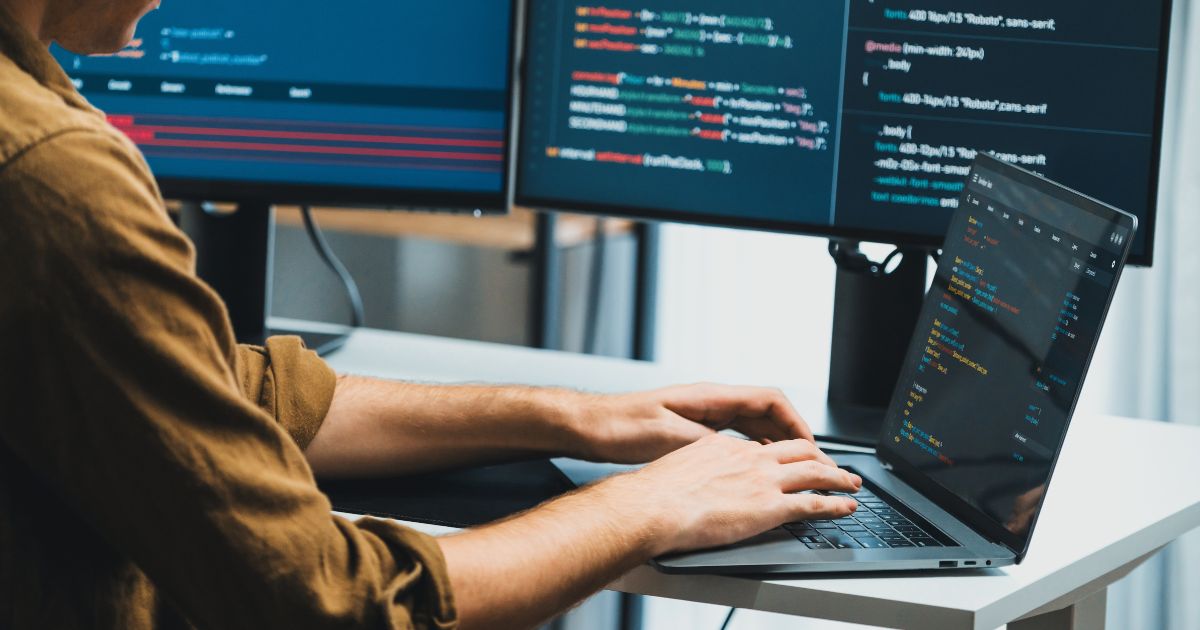 2025, Uma pessoa está trabalhando em um laptop conectado a dois monitores externos, que exibem linhas de código em diferentes cores. O desenvolvedor digita no teclado do laptop, e os três monitores mostram trechos de programação. O ambiente sugere um espaço de trabalho voltado para desenvolvimento de software (brasil, brasscom, treinee, digitização)