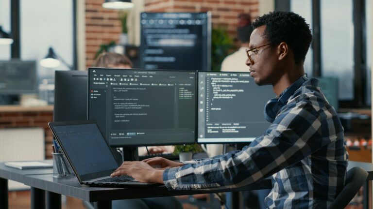 Programador trabalhando em escritório com múltiplos monitores, focado na programação de software (TIC, cargo, reportagens)