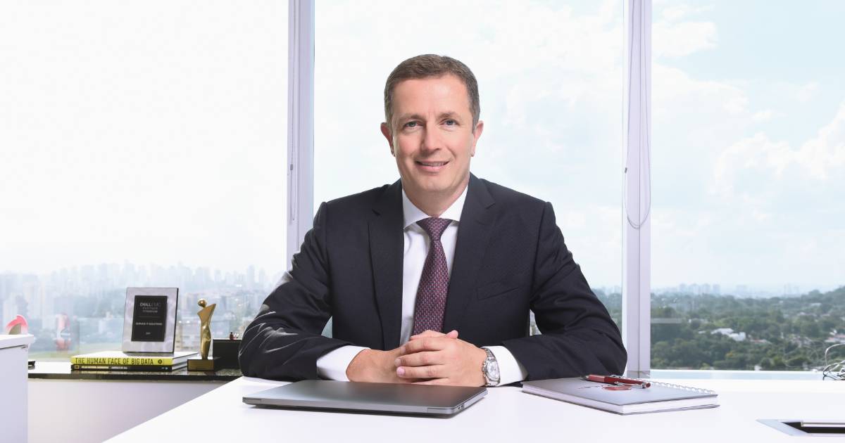 Eduardo Gallo, presidente da Service IT, sentado atrás de uma mesa de escritório com laptop, caderno e caneta, posando para foto com vista panorâmica da cidade ao fundo e prêmios e livros sobre a mesa lateral.
