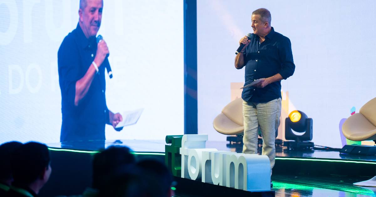 Adelson de Sousa, fundador e presidente do Itaqui, fala ao microfone no palco durante a abertura do IT Forum Praia do Forte, segurando um papel nas mãos, com o logotipo do evento em destaque à frente do palco.