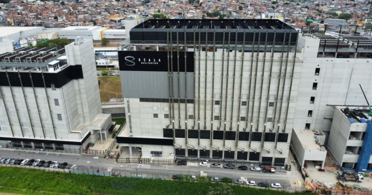 A imagem mostra um grande complexo industrial ou comercial composto por várias estruturas. O edifício principal, localizado ao centro, possui uma placa com os dizeres "SCALA DATA CENTERS". Há diversos carros estacionados ao longo da rua em frente aos prédios, e ao fundo é possível ver uma área urbana densamente povoada.
