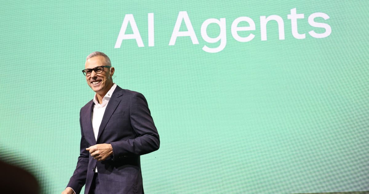 Uma pessoa está em pé diante de um grande painel verde que exibe o texto "AI Agents" em letras brancas. Ela veste um terno escuro com uma camisa branca, transmitindo uma aparência formal e profissional. (Zendesk)