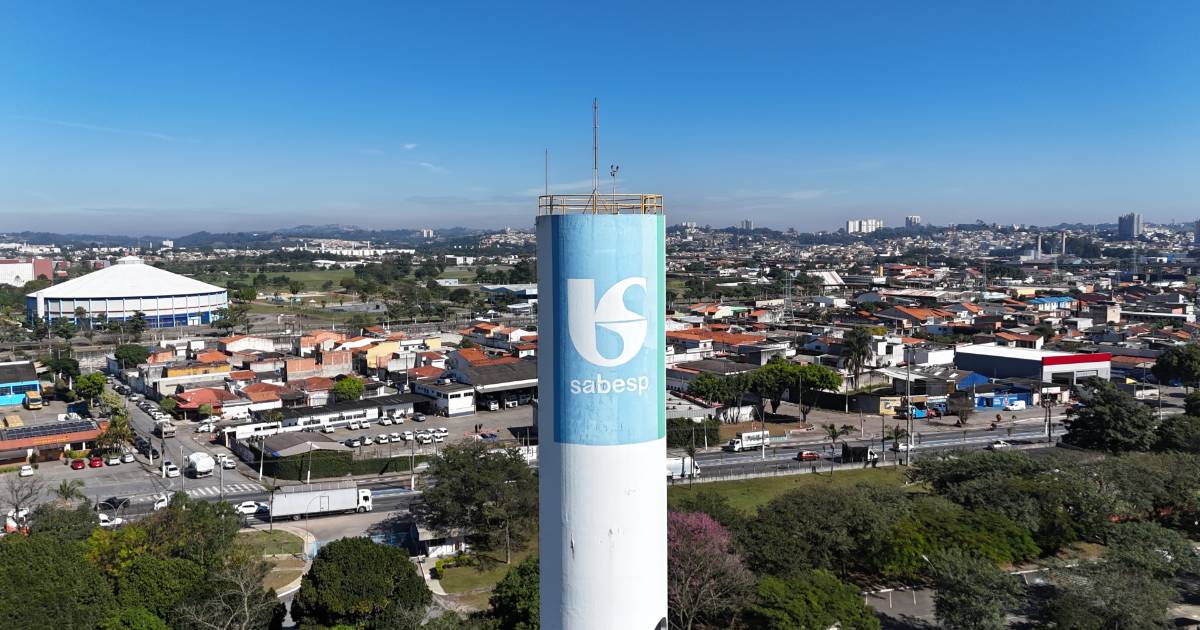Vista aérea de uma região urbana com destaque para um reservatório cilíndrico branco da Sabesp (Companhia de Saneamento Básico do Estado de São Paulo), com o logotipo azul da empresa no centro. Ao fundo, há um bairro com casas, comércios e ruas movimentadas, além de um grande ginásio coberto à esquerda e edifícios espalhados ao longe. O céu está limpo e azul, indicando um dia ensolarado (inteligentes)