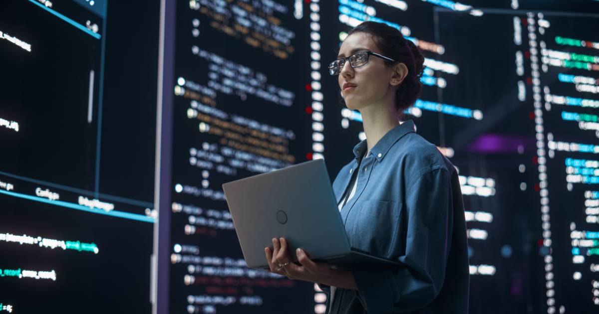 Mulher usando óculos e camisa azul, segurando um notebook, em frente a grandes telas com códigos de programação e dados iluminados (segurança, infraestrutura)