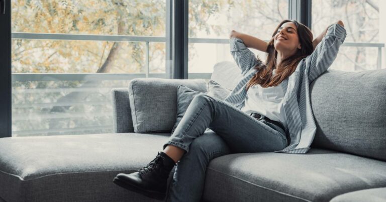 Mulher sentada em um sofá cinza, sorrindo e relaxando com os braços apoiados atrás da cabeça, em frente a uma grande janela com vista para árvores, transmitindo sensação de tranquilidade e bem-estar (vida)