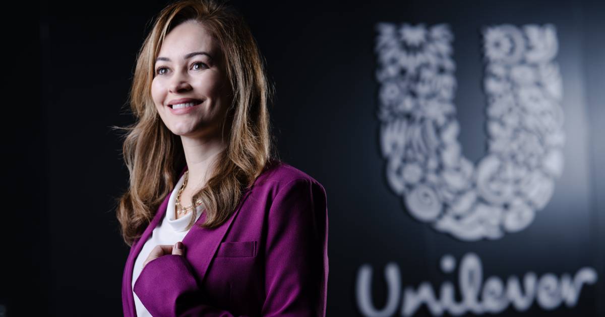 Mulher sorridente com cabelos castanhos claros, vestindo blazer roxo e blusa branca, em frente a uma parede preta com o logotipo da Unilever em destaque. A legenda informa que é Claudia Meira, CIO Latam da Unilever.
