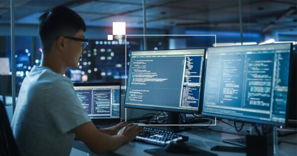 Pessoa desenvolvedora de software trabalhando com AI PCs com múltiplas telas de código em ambiente de escritório à noite. Brasil