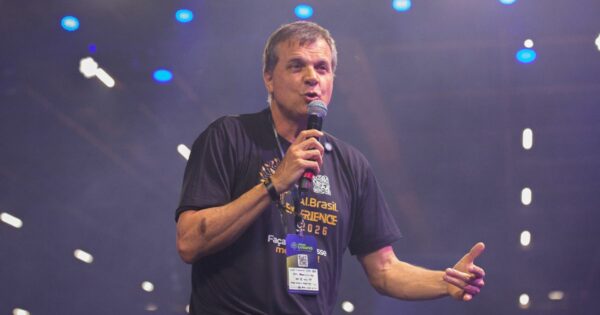 A cena mostra uma pessoa em pé sobre um palco, segurando um microfone na mão direita e gesticulando com a mão esquerda. A pessoa veste uma camiseta preta com texto em amarelo e branco que inclui as palavras “AI Brasil Experience 2026” e um crachá pendurado no pescoço com QR code e logotipos de patrocinadores. O fundo está iluminado com luzes roxas e azuis, criando um efeito de evento tecnológico ou conferência, com vários pontos de luz desfocados ao fundo, sugerindo um ambiente amplo e moderno. (IA)