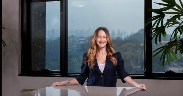 A imagem mostra uma pessoa em pé atrás de uma mesa, em um ambiente de escritório. Ela veste um blazer escuro sobre uma blusa clara e tem cabelo longo e ondulado. Ao fundo, há grandes janelas que revelam uma paisagem urbana com áreas verdes, além de plantas internas visíveis à direita. A composição transmite um contexto profissional, ideal para ilustrar temas relacionados a negócios ou ambientes corporativos. (Gisselle, liderança)