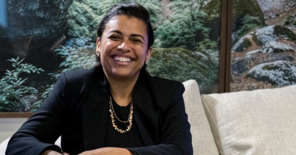 Mulher sorridente sentada em um sofá claro, usando blazer preto e colar dourado, com um painel de paisagem natural ao fundo; junto à imagem aparece o texto “Noéle Gomes, pesquisadora”. (filosofia)