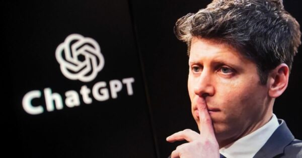 A foto mostra uma pessoa em primeiro plano, com fundo escuro onde aparece o logotipo do ChatGPT em destaque. O logotipo inclui um ícone estilizado à esquerda e o texto “ChatGPT” em letras brancas à direita. A cena sugere um ambiente de apresentação ou evento relacionado à tecnologia e inteligência artificial. (OpenAI)