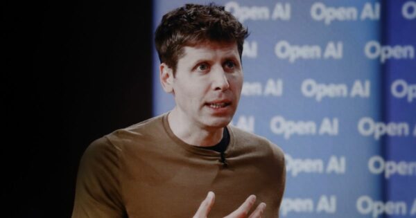 A cena mostra um grande painel branco com a palavra “OPENAI” escrita em letras maiúsculas pretas, ocupando a maior parte do fundo. Em frente ao painel, há uma pessoa em pé, segurando um microfone, sugerindo que está fazendo uma apresentação ou discurso. No primeiro plano, aparecem silhuetas desfocadas de outras pessoas, indicando que a imagem foi capturada durante um evento ou conferência. A iluminação destaca fortemente o painel, criando contraste com as figuras sombreadas à frente, reforçando um ambiente moderno e tecnológico.
