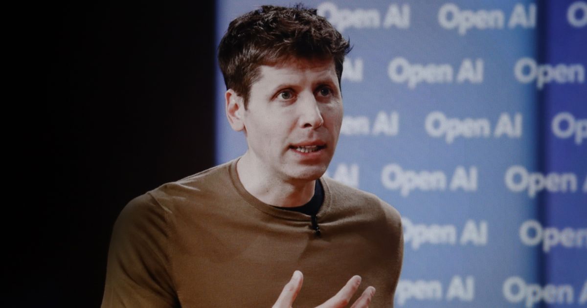 A cena mostra um grande painel branco com a palavra “OPENAI” escrita em letras maiúsculas pretas, ocupando a maior parte do fundo. Em frente ao painel, há uma pessoa em pé, segurando um microfone, sugerindo que está fazendo uma apresentação ou discurso. No primeiro plano, aparecem silhuetas desfocadas de outras pessoas, indicando que a imagem foi capturada durante um evento ou conferência. A iluminação destaca fortemente o painel, criando contraste com as figuras sombreadas à frente, reforçando um ambiente moderno e tecnológico.