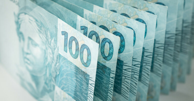 Várias cédulas de 100 reais alinhadas em sequência, mostrando detalhes em azul da nota brasileira e a figura da efígie da República parcialmente visível