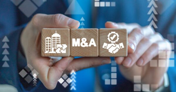 Duas mãos seguram três blocos de madeira alinhados horizontalmente. O bloco central exibe a sigla “M&A”, enquanto os blocos laterais apresentam ícones de edifícios corporativos e um aperto de mãos com símbolo de verificação. Ao fundo, há elementos gráficos abstratos em tons de azul, com setas e formas geométricas, sugerindo crescimento, integração empresarial e processos de fusões e aquisições em um contexto corporativo e financeiro. (M&A)