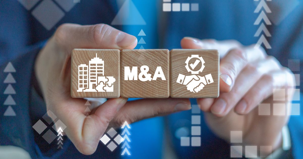Duas mãos seguram três blocos de madeira alinhados horizontalmente. O bloco central exibe a sigla “M&A”, enquanto os blocos laterais apresentam ícones de edifícios corporativos e um aperto de mãos com símbolo de verificação. Ao fundo, há elementos gráficos abstratos em tons de azul, com setas e formas geométricas, sugerindo crescimento, integração empresarial e processos de fusões e aquisições em um contexto corporativo e financeiro. (M&A)