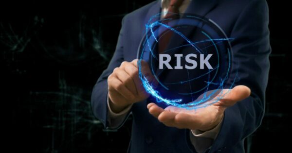 Pessoa vestindo terno segura um holograma azul em forma de esfera, com linhas de energia brilhantes ao redor. No centro da esfera aparece a palavra “RISK” (riscos). A cena tem aparência tecnológica e futurista, com o fundo escuro destacando o efeito luminoso do holograma.