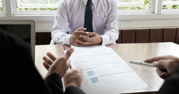 A imagem mostra uma cena típica de entrevista ou reunião profissional em um ambiente corporativo. Detalhes principais: Primeiro plano: Duas mãos segurando um currículo ou documento impresso, com texto visível em formato de lista, indicando que é provavelmente um CV ou proposta. Outra mão segura uma caneta, sugerindo anotações ou avaliação. Centro da imagem: Pessoa vestindo camisa social branca e gravata listrada azul, com as mãos entrelaçadas sobre a mesa, postura formal e atenta. Ambiente: Mesa de madeira clara, com um laptop parcialmente visível à esquerda. Fundo com janelas amplas, permitindo entrada de luz natural, e plantas do lado externo, criando um clima profissional e acolhedor. Essa imagem transmite claramente processos seletivos, entrevistas de emprego ou reuniões de negócios.