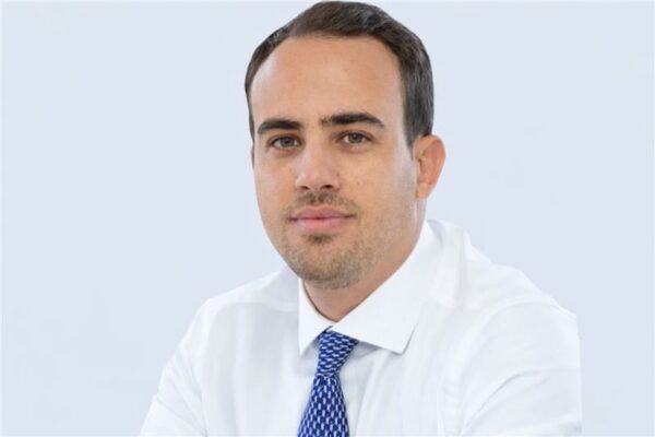 Pessoa vestindo uma camisa social branca e uma gravata azul com padrão discreto, sentada diante de um fundo claro e uniforme. A pose é formal, sugerindo um retrato profissional ou corporativo. (telemedicina)