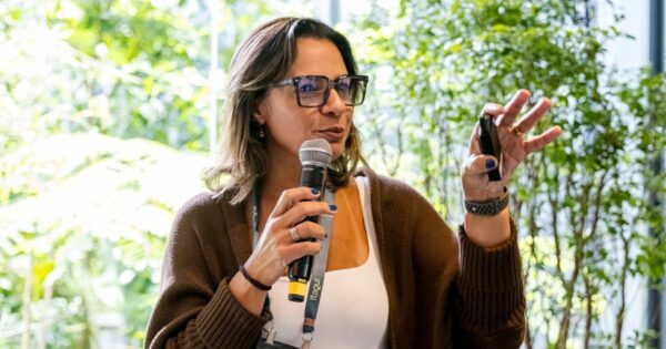 Uma pessoa está falando em um ambiente iluminado por luz natural, segurando um microfone em uma mão e um controle remoto de apresentação na outra. Ela veste um cardigã marrom sobre uma blusa clara e usa óculos. Ao fundo, há plantas verdes que compõem um cenário natural e suave. (mudança)