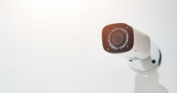 Uma câmera de segurança fixa está posicionada em uma parede clara, voltada para frente. O equipamento possui corpo branco e lente central cercada por LEDs infravermelhos, sugerindo capacidade de gravação mesmo em baixa luminosidade. A iluminação suave no canto superior esquerdo cria um clima minimalista e clean. (vigilância)