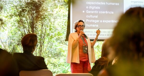 Uma pessoa em pé, usando roupas claras e segurando um microfone, faz uma apresentação diante de um pequeno público. Ela está posicionada entre um ambiente externo com vegetação ao fundo e um telão que exibe texto relacionado a capacidades humanas, coerência cultural e experiência humana. O público aparece sentado em primeiro plano, observando a apresentação. (governança)
