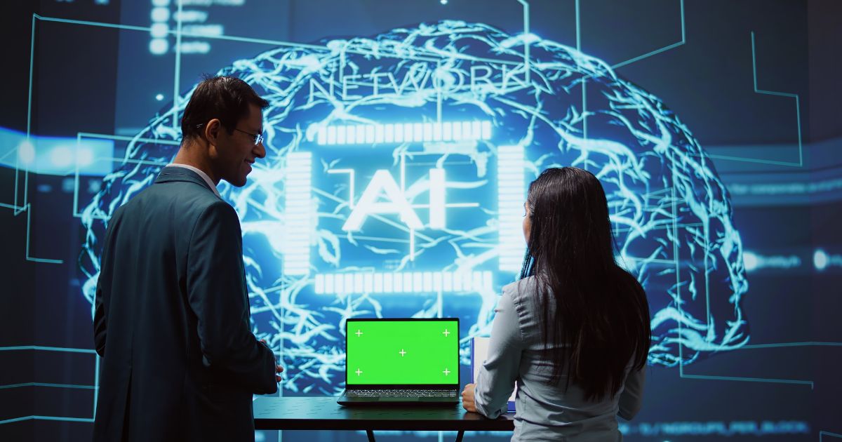 Duas pessoas observam uma grande projeção digital que exibe a ilustração de um cérebro formado por circuitos e linhas luminosas em azul, com a palavra “AI” ao centro. À frente delas, há um notebook aberto sobre uma mesa, com a tela em verde sólido, sugerindo uso de chroma key. O ambiente tem aparência tecnológica e imersiva, como uma demonstração ou apresentação de sistemas de inteligência artificial. (seguradora)