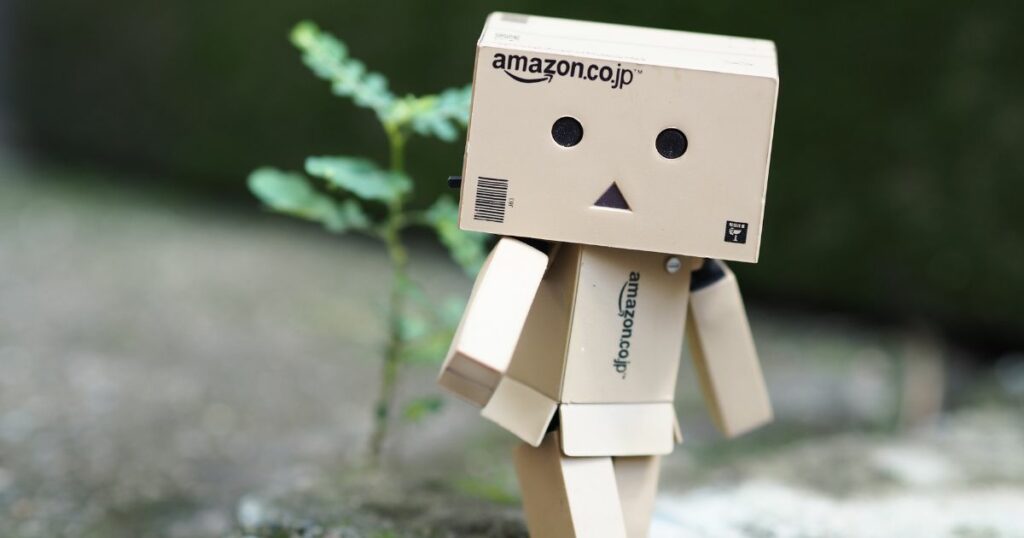 Uma pequena figura feita de caixas de papelão está posicionada ao ar livre, sobre uma superfície de concreto desfocada. A cabeça é uma caixa com o logotipo “amazon.co.jp” impresso na frente, e o rosto é composto por dois círculos pretos como olhos e um pequeno triângulo preto no centro como boca. O corpo e os membros também são formados por partes retangulares de papelão articuladas. A figura está levemente inclinada para o lado, como se estivesse dando um passo. Ao fundo, há um pequeno broto verde crescendo no solo e uma área desfocada em tons de verde e cinza. (Robos Amazon)