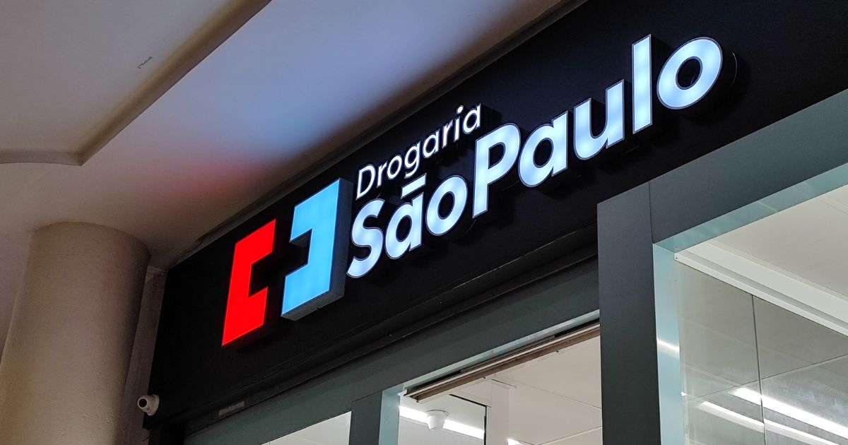 A imagem mostra a fachada de uma loja da Drogaria São Paulo vista de um ângulo levemente inclinado para cima. O letreiro luminoso tem as palavras “Drogaria São Paulo” escritas em letras brancas e brilhantes. À esquerda do texto há um símbolo composto por dois quadrados: um vermelho e outro azul‑claro, formando um design geométrico. A fachada é preta e, logo abaixo do letreiro, é possível ver parte da entrada da loja, com portas de vidro e iluminação interna branca. (Grupo DPSP)